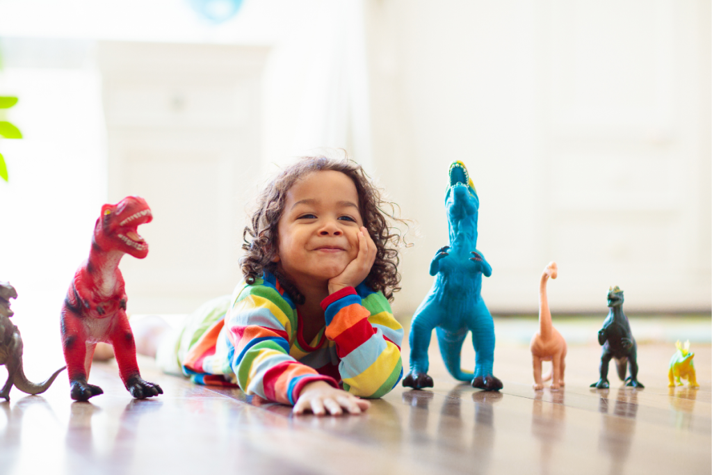 A smiling ND child in a rainbow shirt proudly surrounded by dinosaur toys, a perfect example of celebrating ND child passions and embracing unique interests.
