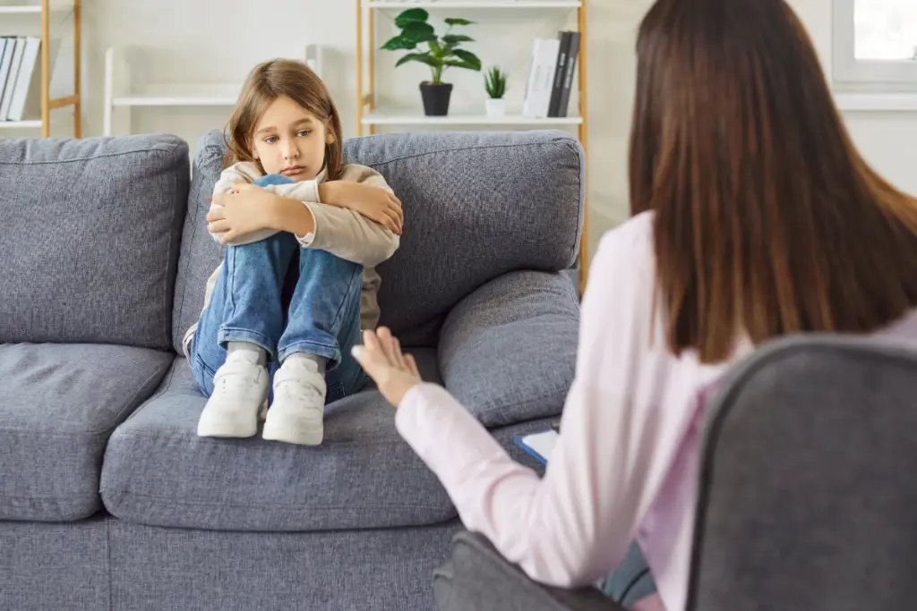 A young girl sits curled up on a couch with a worried expression while an adult gently speaks to her—illustrating how hidden stressors can quietly overwhelm neurodivergent children even when they appear calm on the outside.