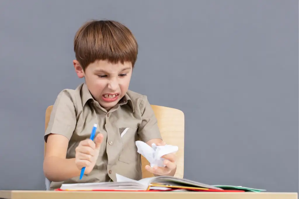 A young boy sits at a desk clenching a pen and crumpling paper in frustration, showing how hidden stressors can overwhelm neurodivergent children during tasks that seem simple on the surface.