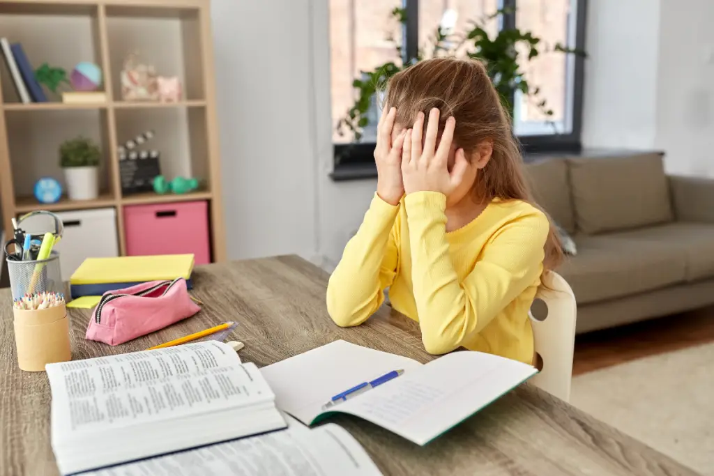 A young girl sits at a desk with her head in her hands, surrounded by school books and supplies, showing how hidden stressors during homework or learning tasks can overwhelm neurodivergent children.