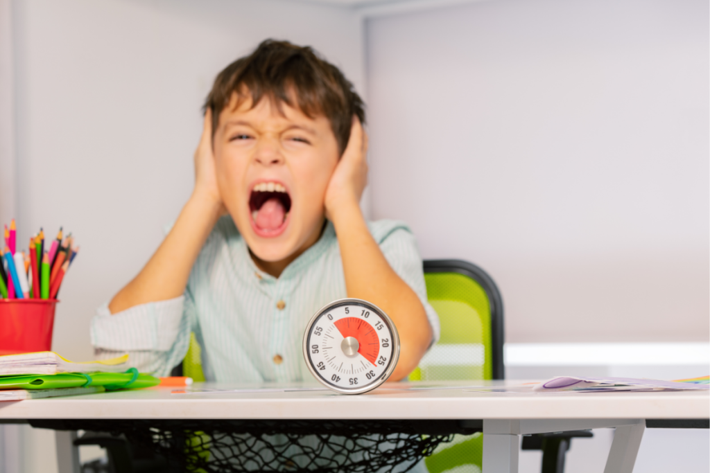 A young boy sits at a desk with his hands over his ears and his mouth open in distress, with a visual timer in front of him—illustrating the sensory overload and emotional strain that often lead to ND masking in neurodivergent children.