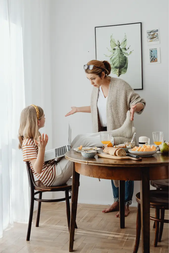 A mother and child sit at a kitchen table during a tense conversation, with the child holding a laptop and both raising their hands in frustration—capturing the everyday misunderstandings that can happen when ND masking hides a neurodivergent child’s true needs.
