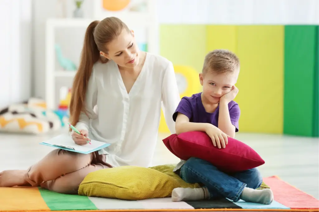 A young boy sits on the floor hugging a pillow and smiling shyly while a clinician observes and takes notes beside him—illustrating how ND masking can make neurodivergent children appear calm or compliant even when they’re working hard to hide their true feelings.